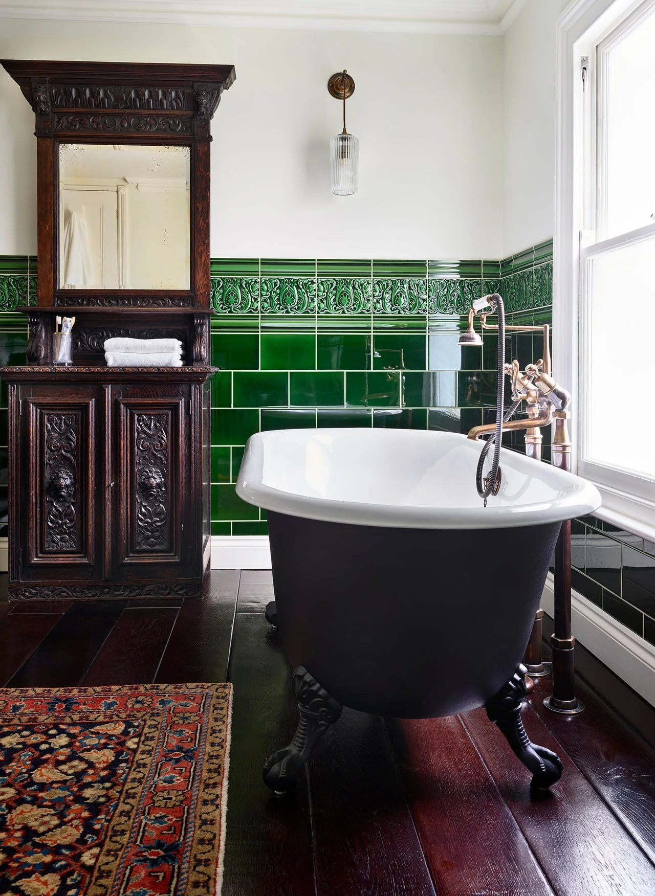 Period-style bathroom with deep green glazed tile, black roll-top bath, dark carved vanity and patterned rug